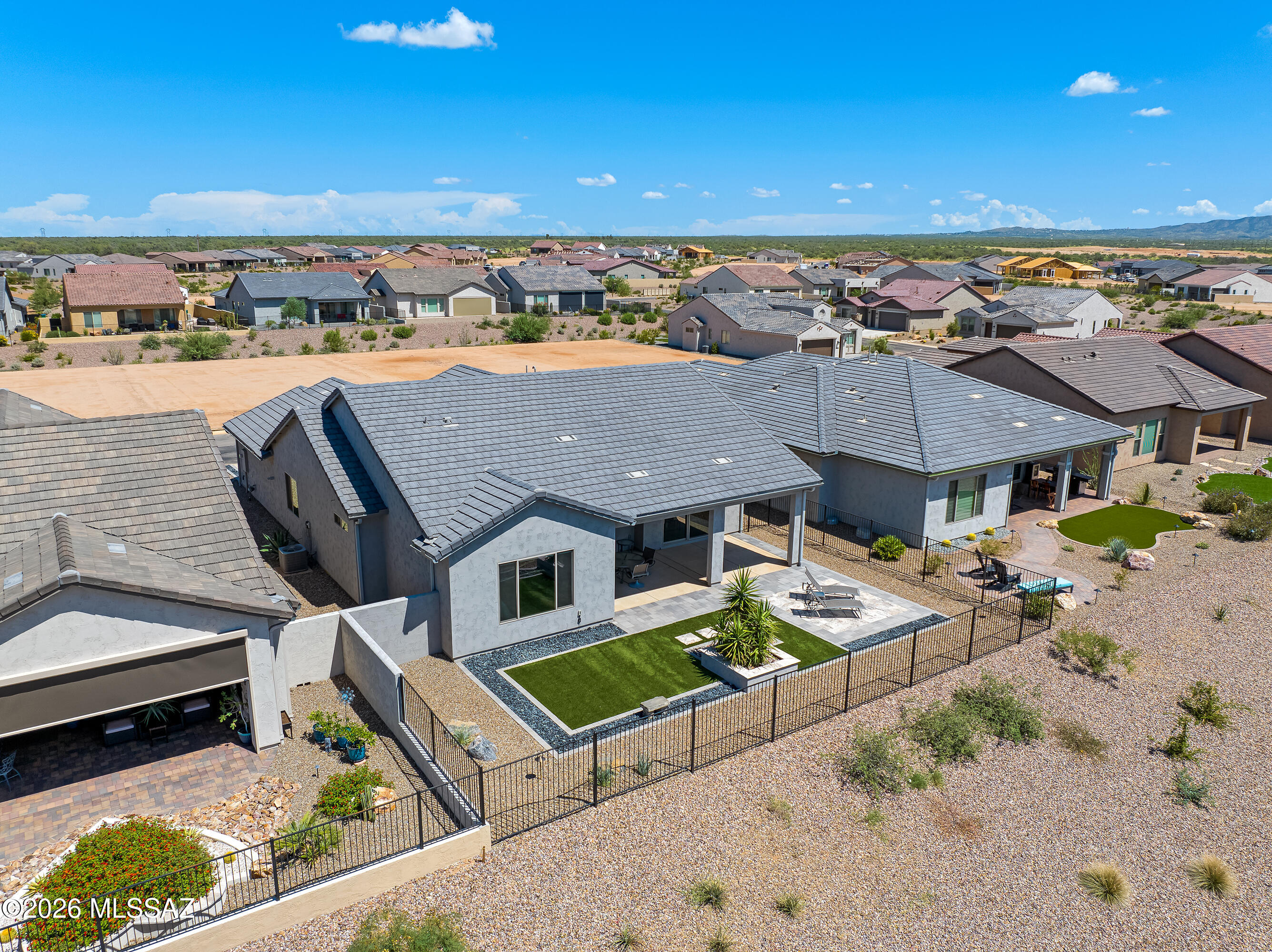 Aerial view of a gray-roofed home with a fenced backyard, artificial turf, and covered patio.