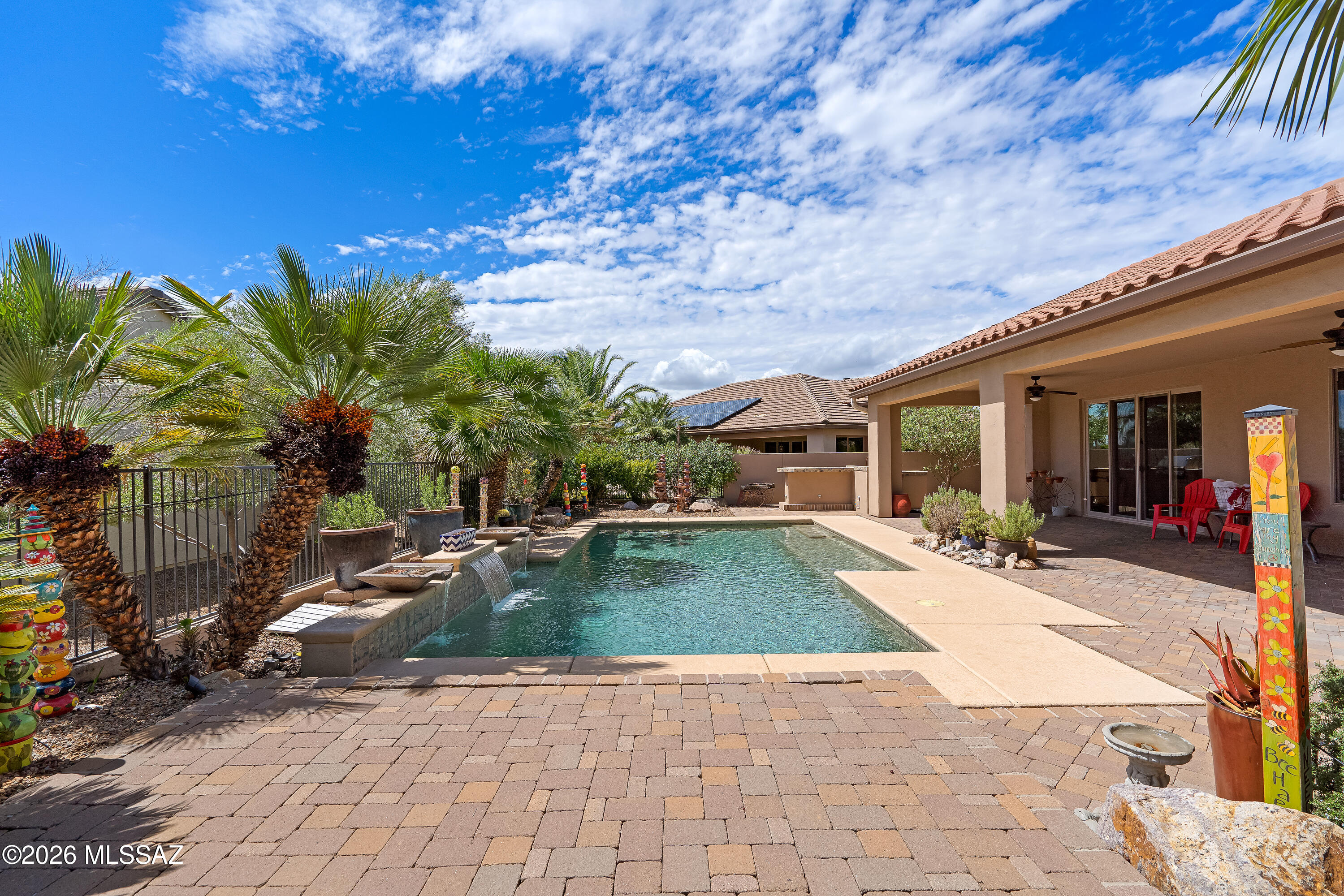 Backyard pool beside a covered patio, with palm trees and a raised waterfall feature.