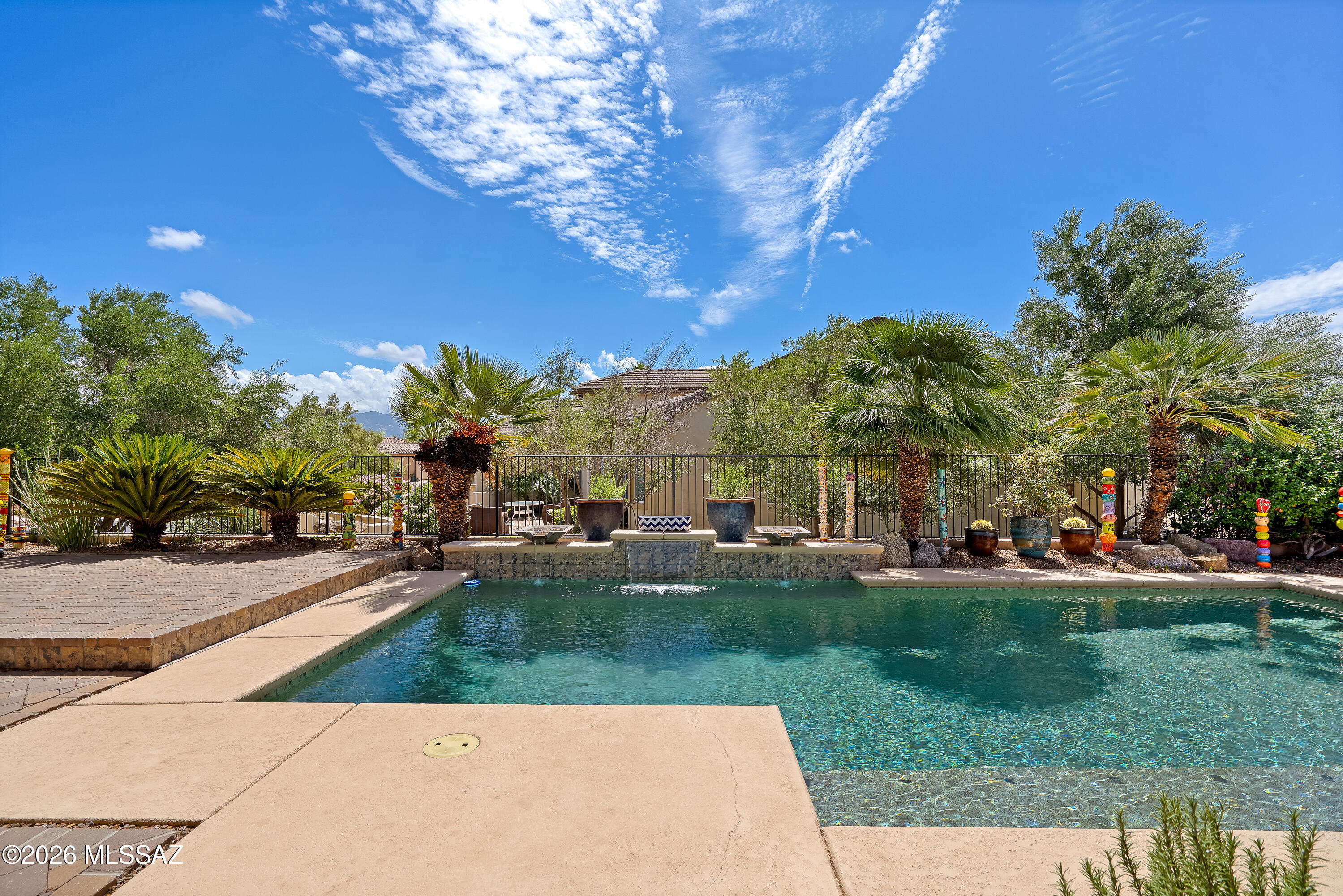 Backyard pool with raised stone water feature, palm trees, and colorful garden ornaments.