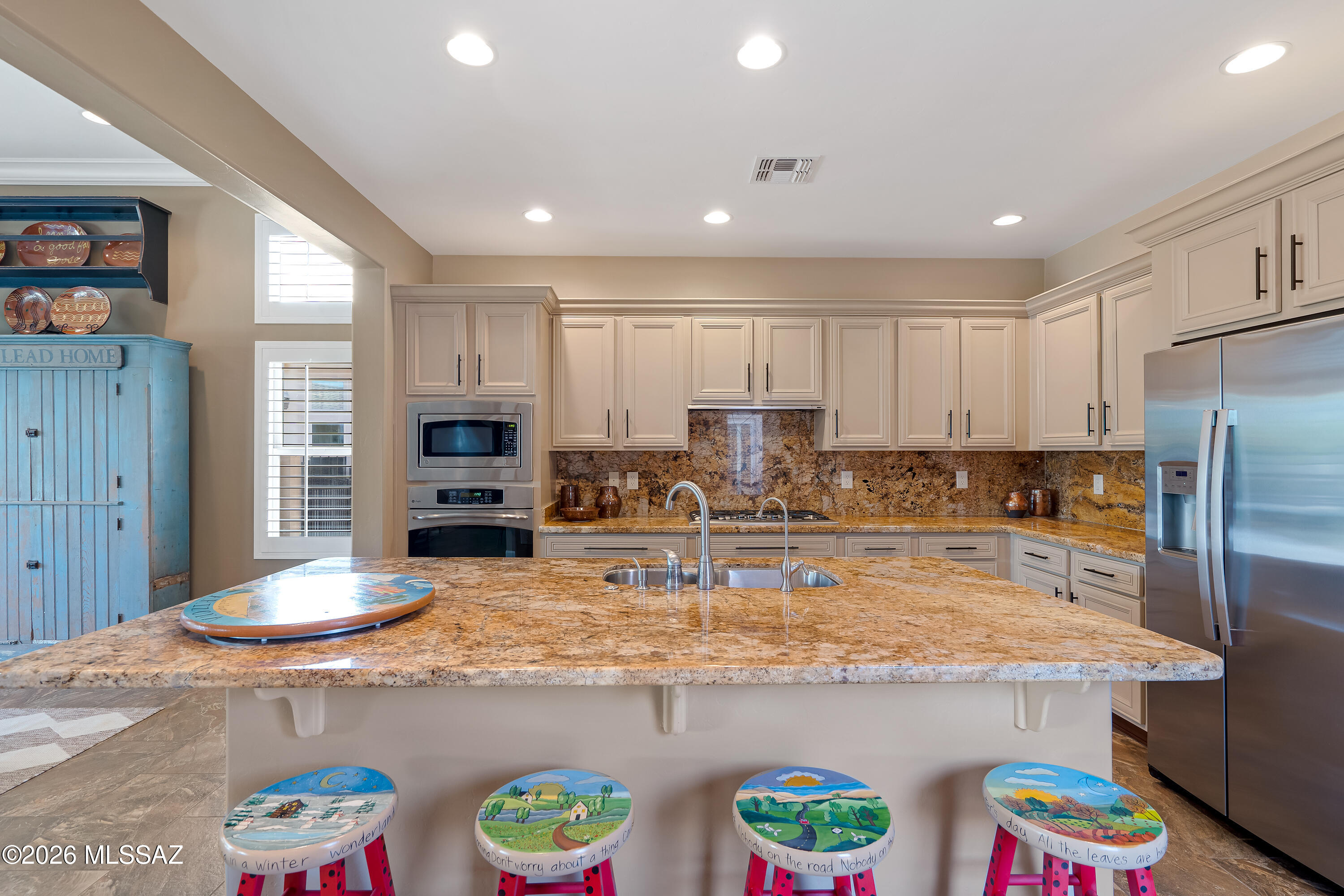 Kitchen with a large stone island, cream cabinets, stainless appliances, and four colorful painted stools.