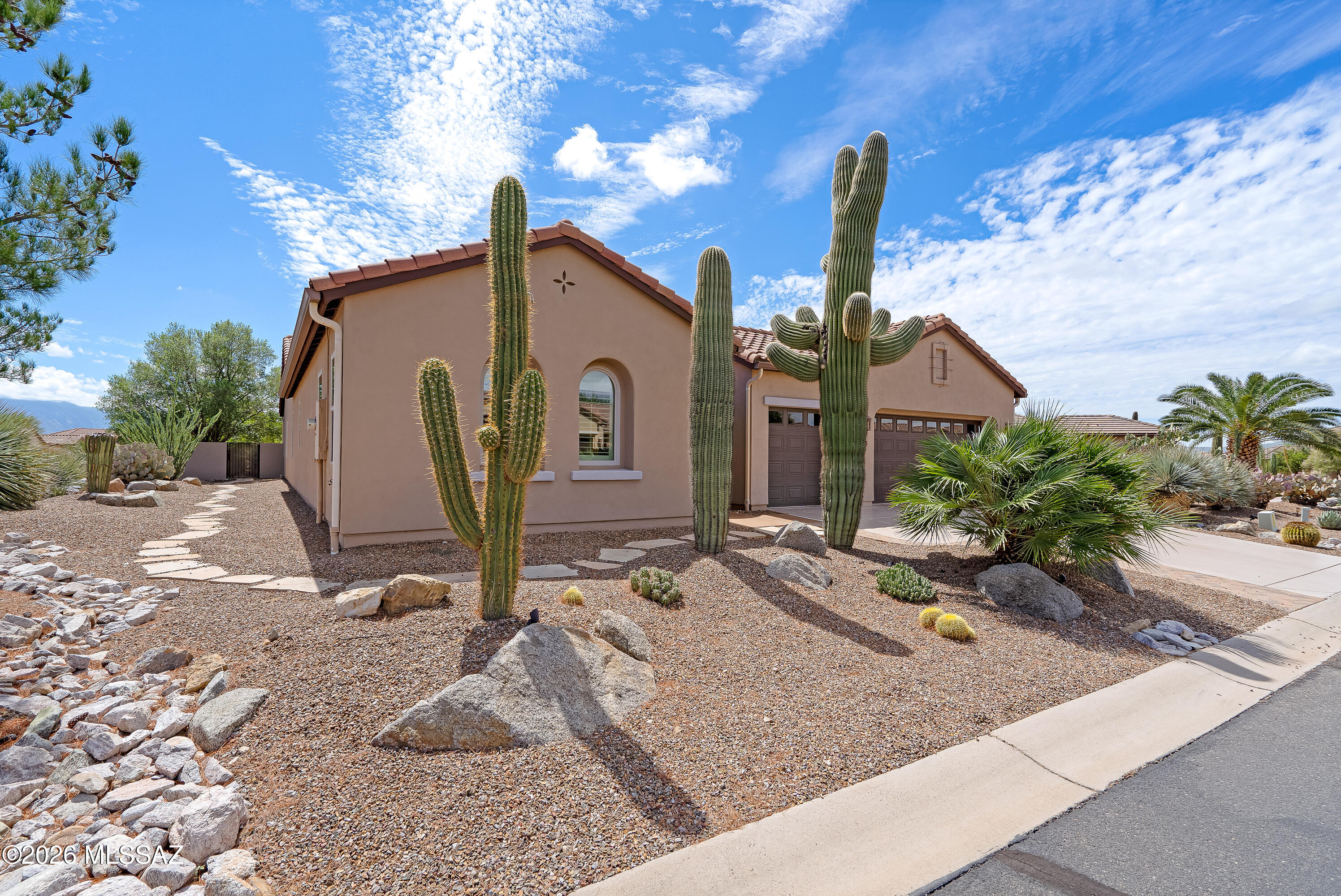 Tan stucco home with tiled roof and garage, framed by tall saguaros and desert rock landscaping.
