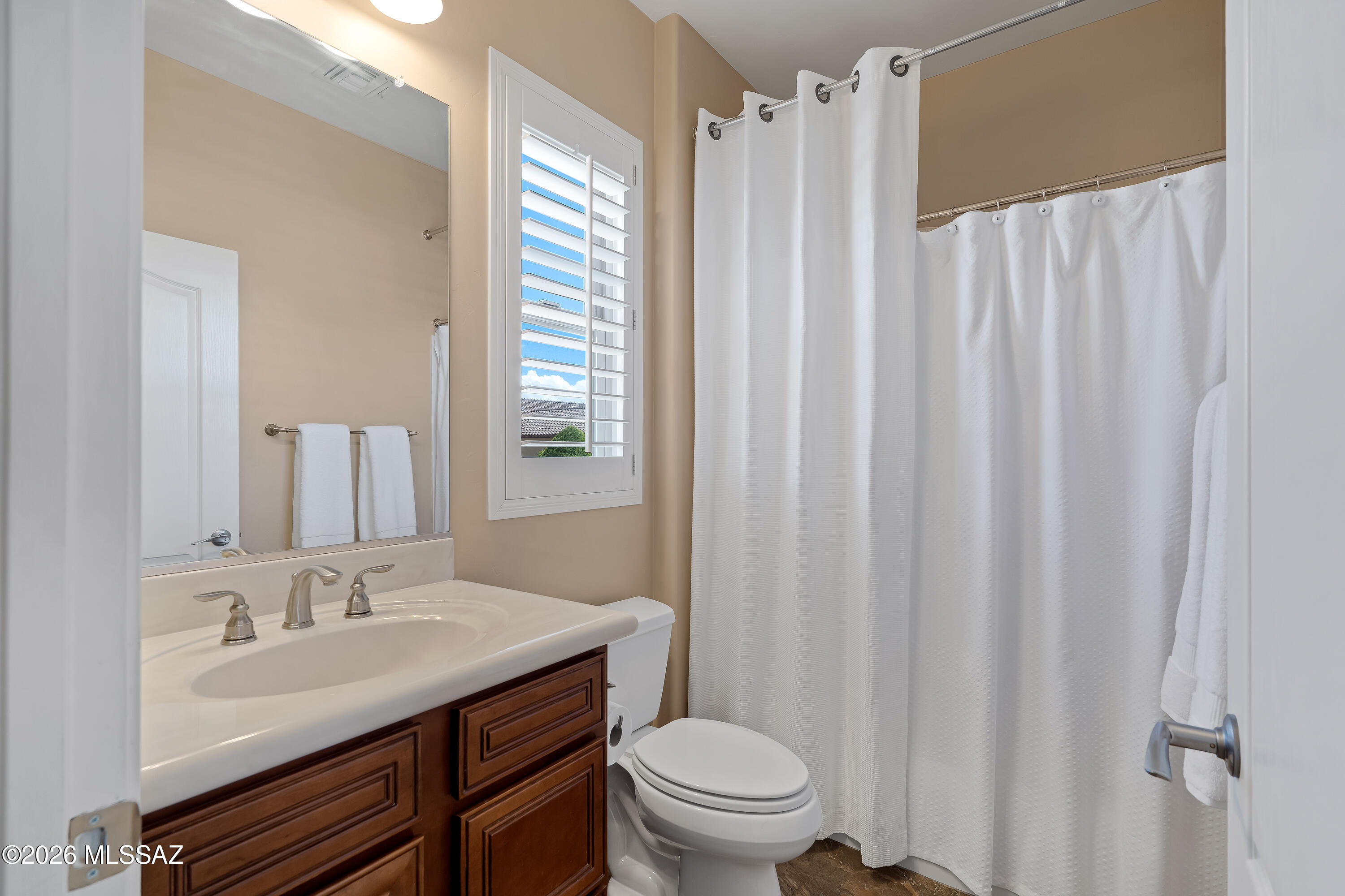 Bathroom with a dark wood vanity, white toilet, curtained tub, and shuttered window.