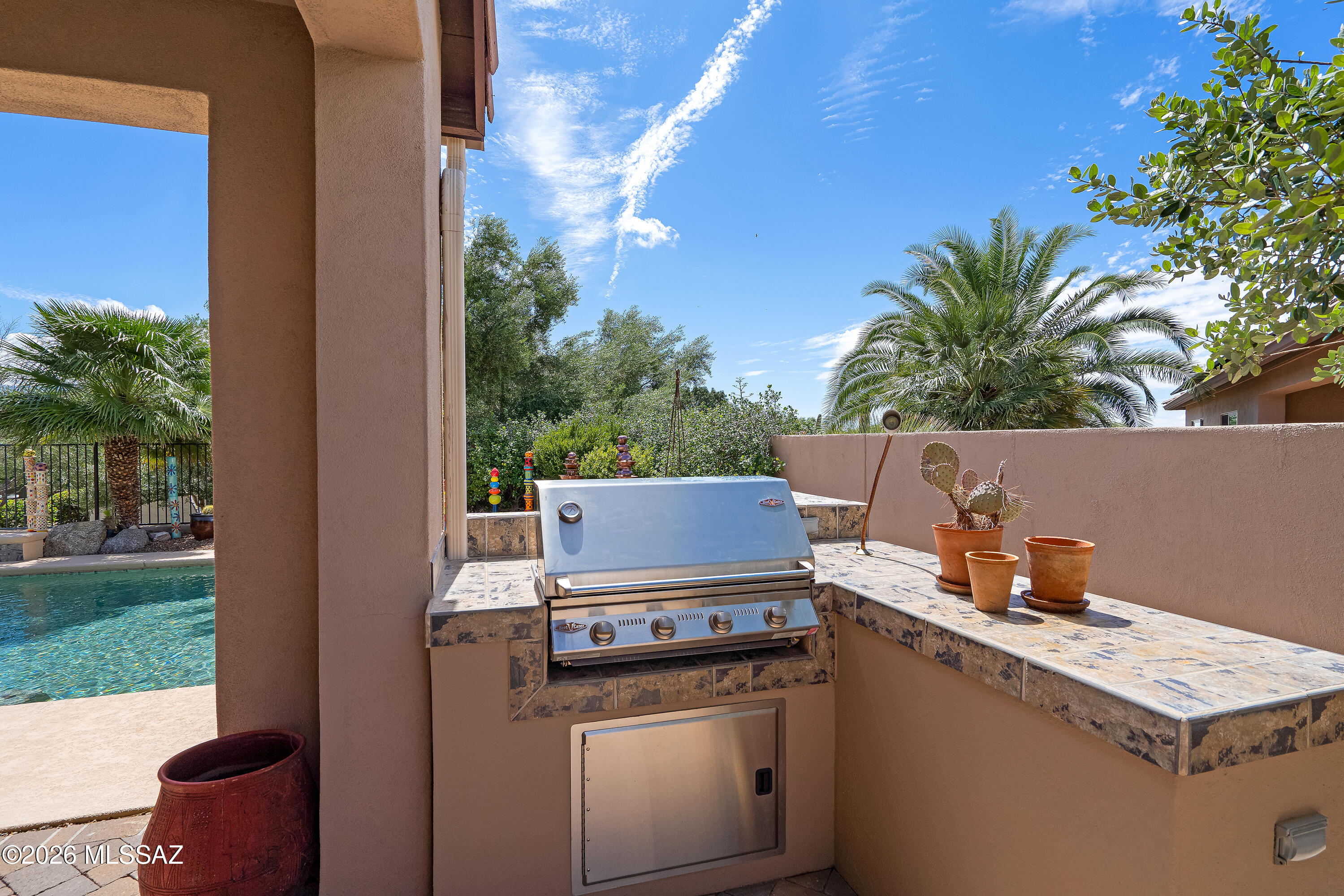 Built-in patio grill beside a swimming pool, with tiled counters and potted cactus.