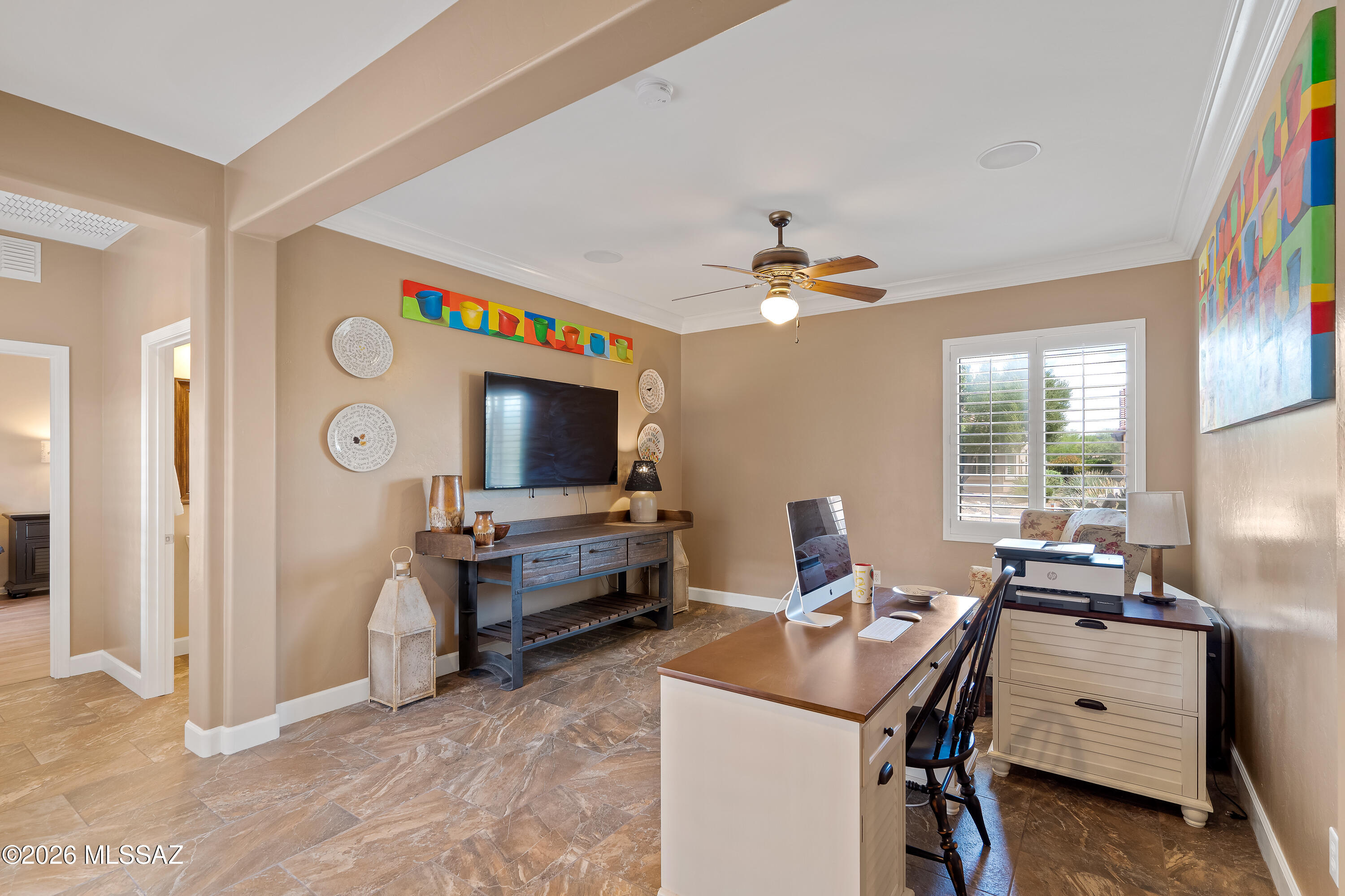 Furnished home office with a large desk, wall-mounted television, colorful artwork, and shuttered window.