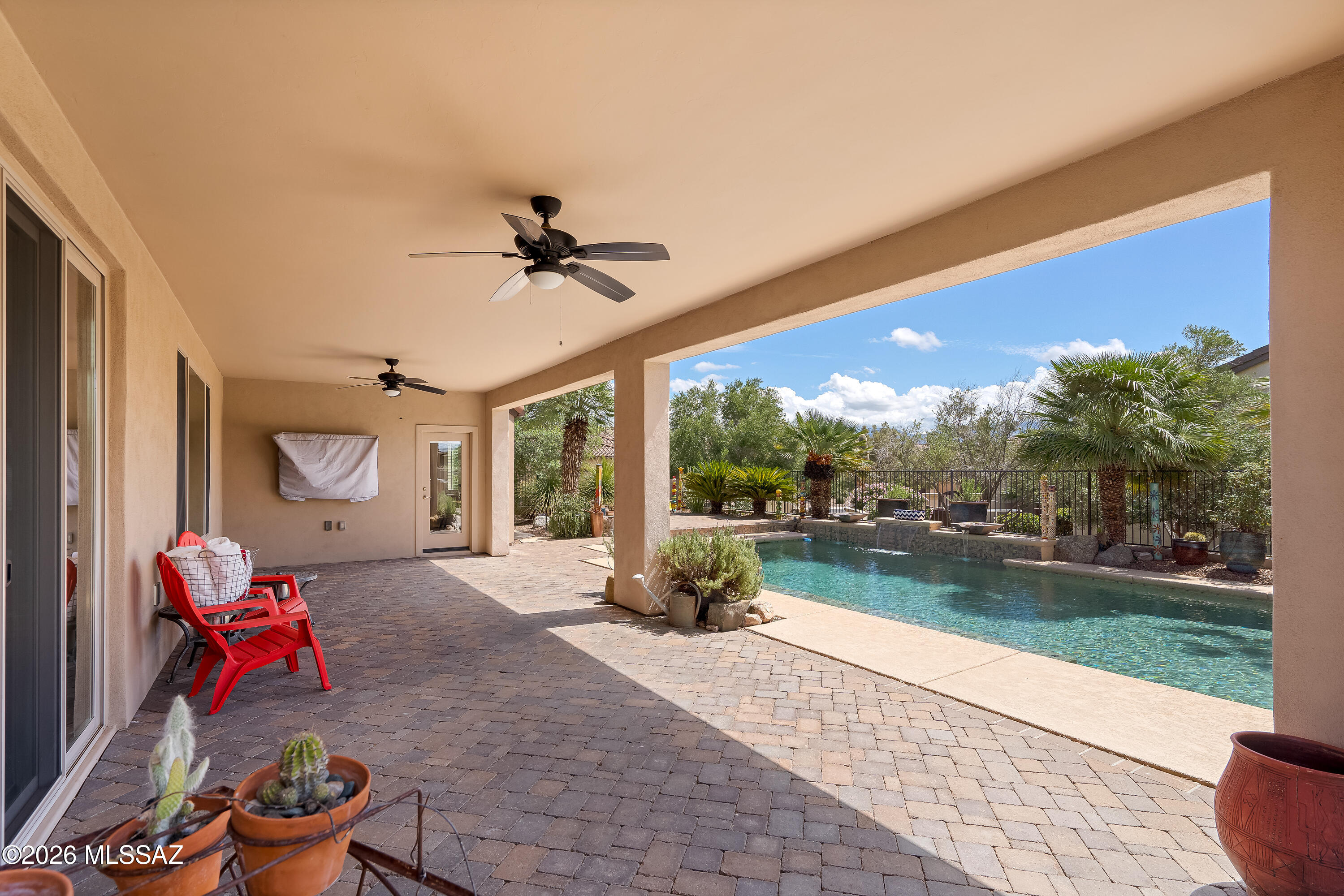 Covered patio with ceiling fans, red chairs, and a swimming pool amid desert landscaping.