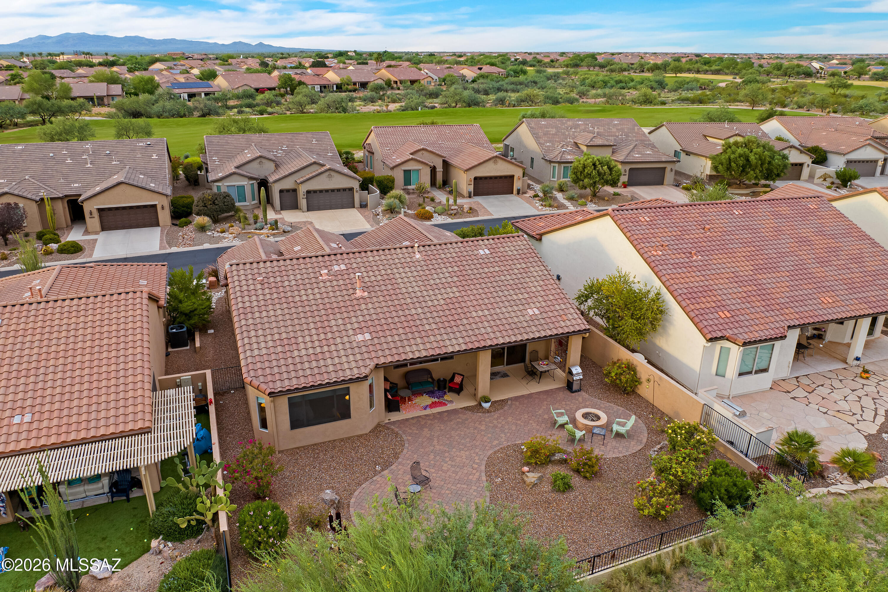 Aerial view of a desert home with a covered patio, paved backyard, circular fire pit, and distant mountain views.