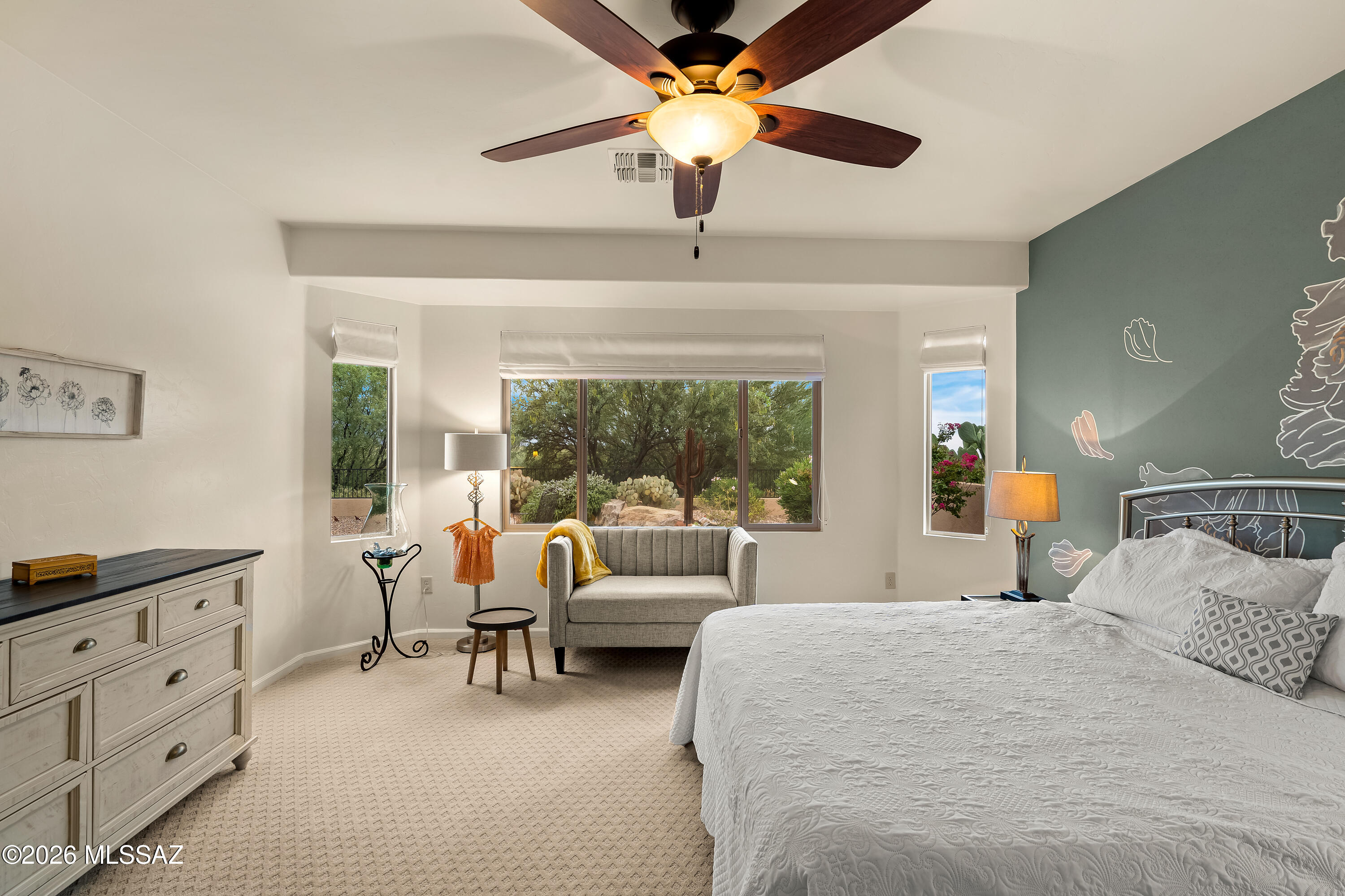 Bedroom with a white quilted bed, gray loveseat, and windows overlooking desert landscaping.