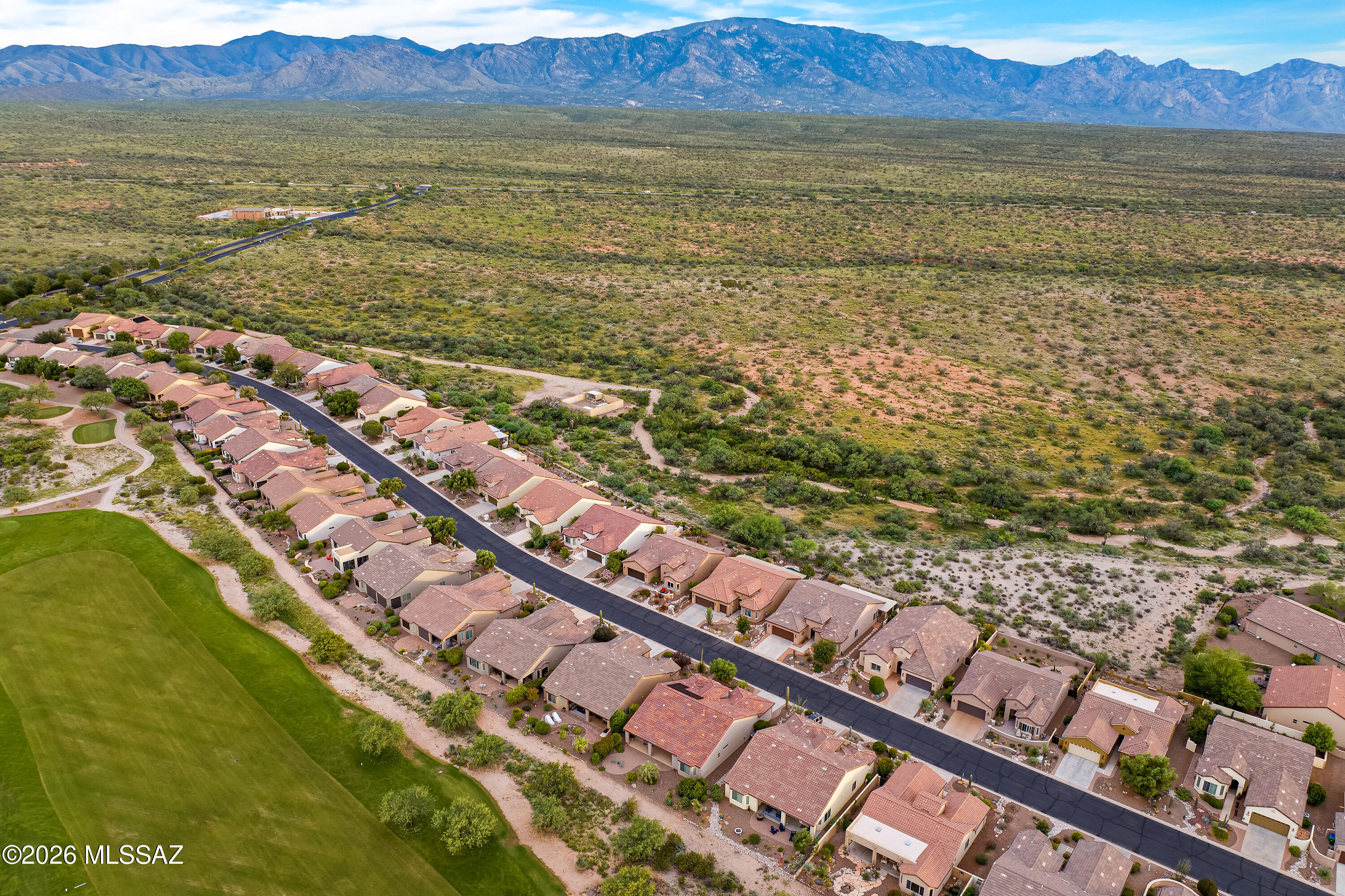 Aerial view of homes bordering a golf course and open desert, with mountains in the distance.