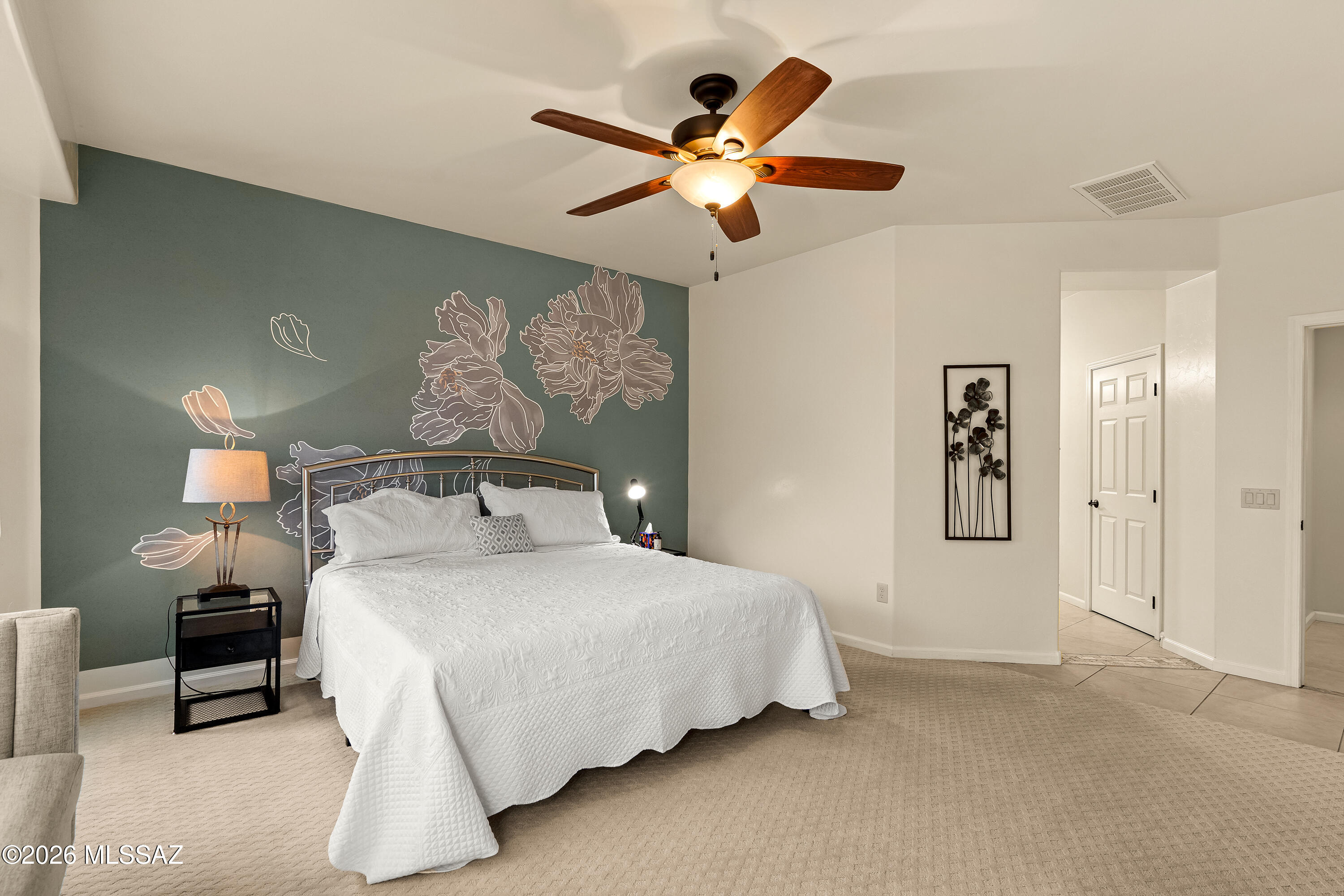Furnished bedroom with a white quilted bed, floral mural, and wood-blade ceiling fan.