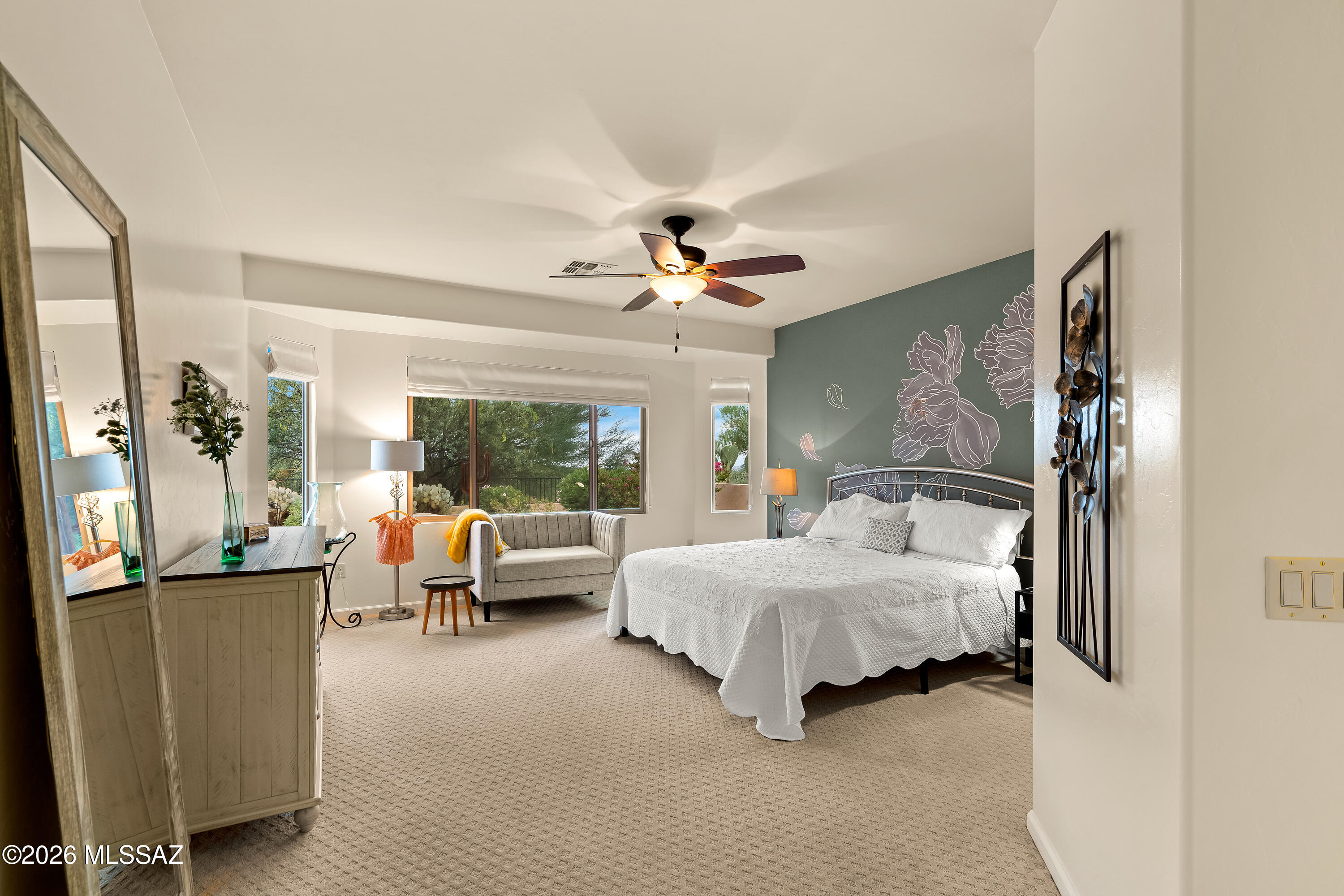 Furnished bedroom with a white bed, gray loveseat, and floral accent wall beside desert-view windows.