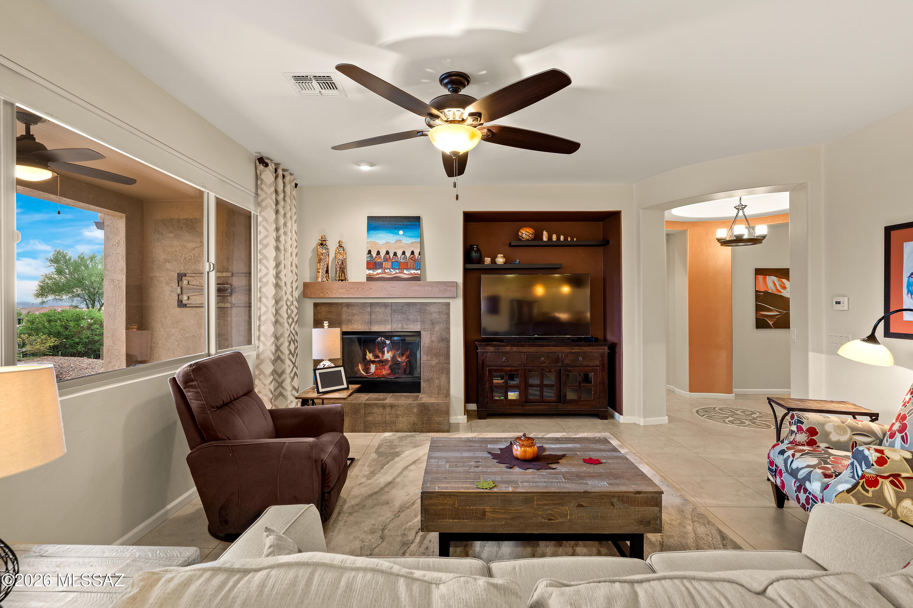 Furnished living room with a tiled fireplace, built-in TV alcove, and large windows overlooking desert landscaping.