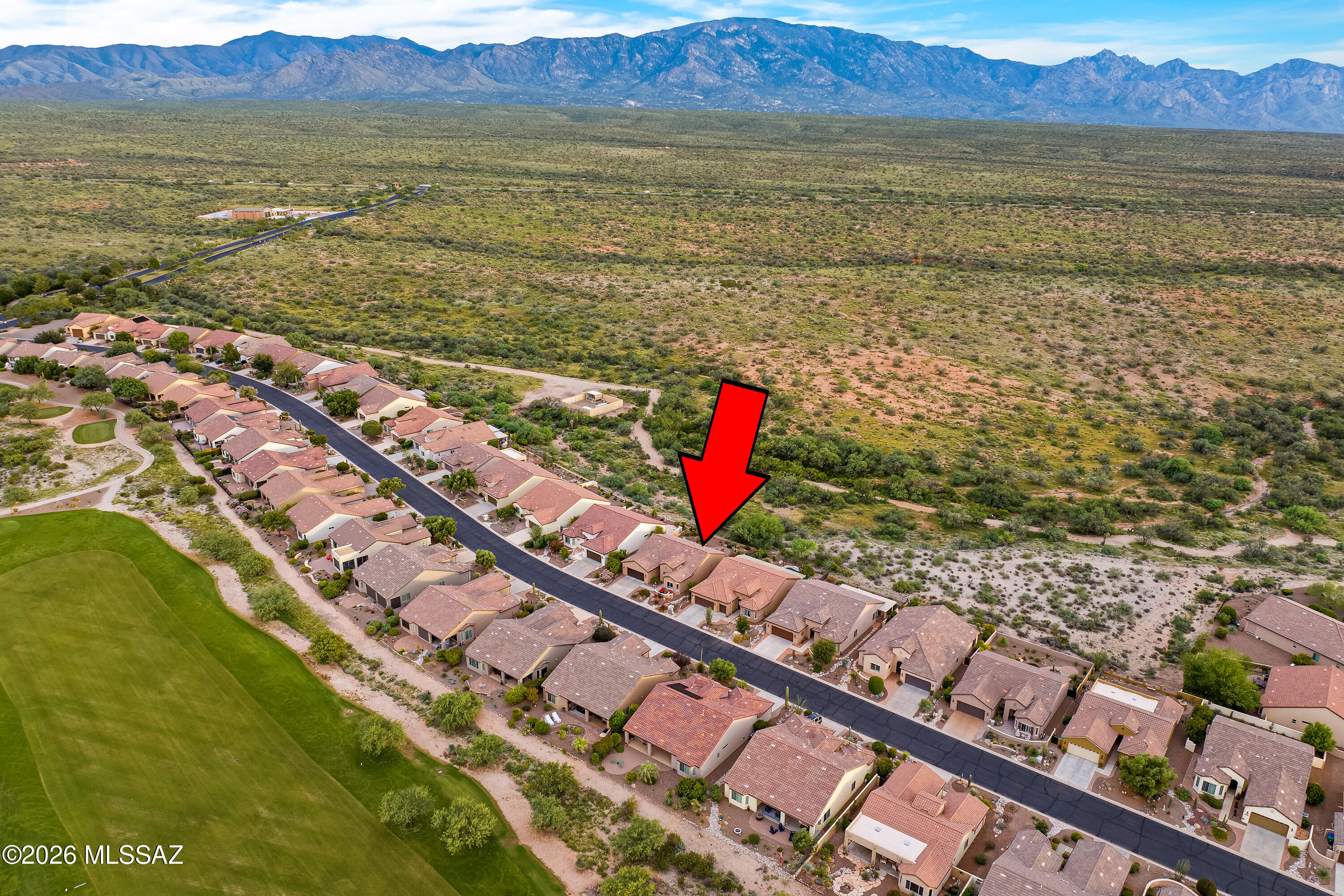 Aerial view of a home marked by a red arrow, beside a golf course with desert and mountains beyond.