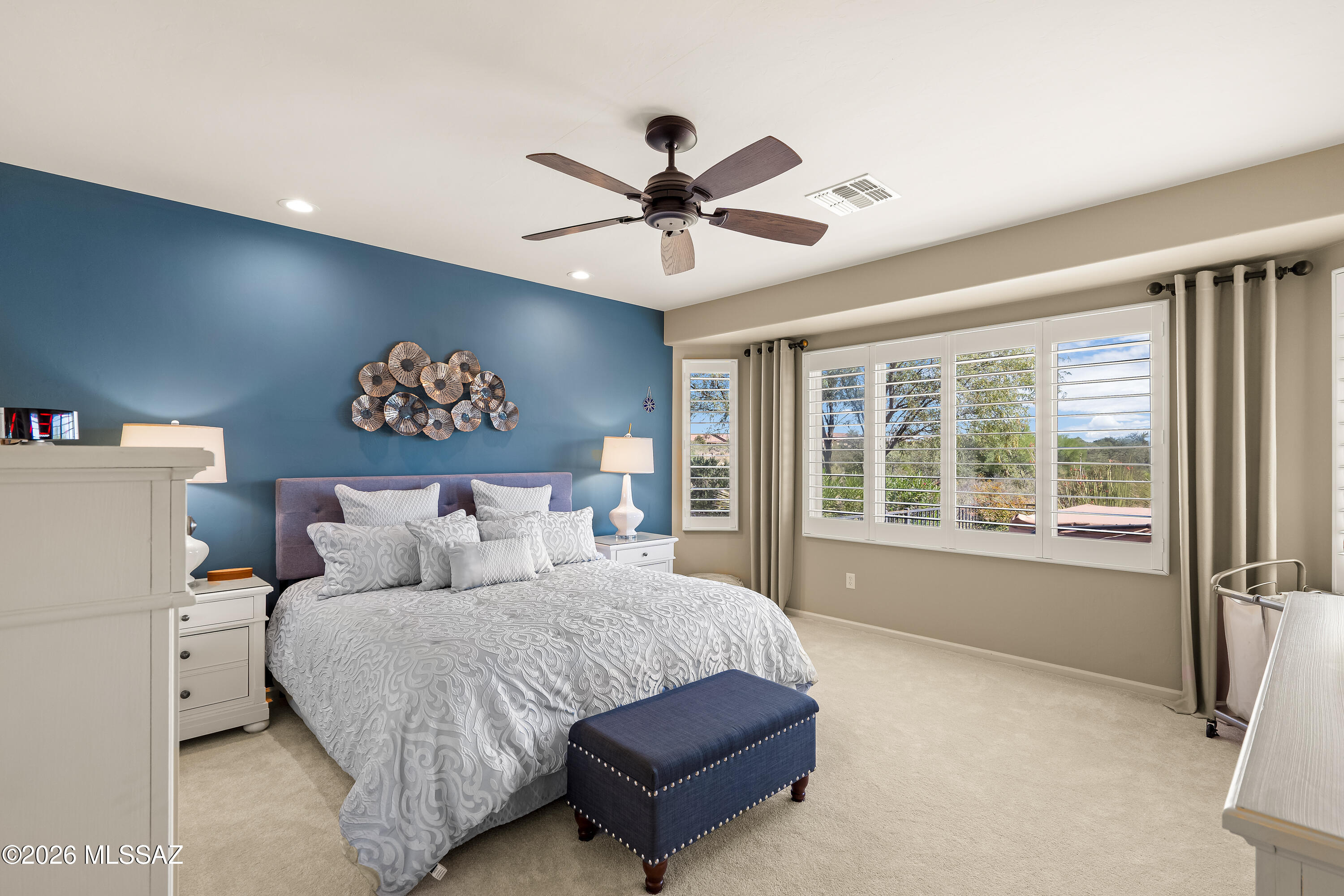 Furnished bedroom with patterned white bedding, a blue accent wall, and large shuttered windows.