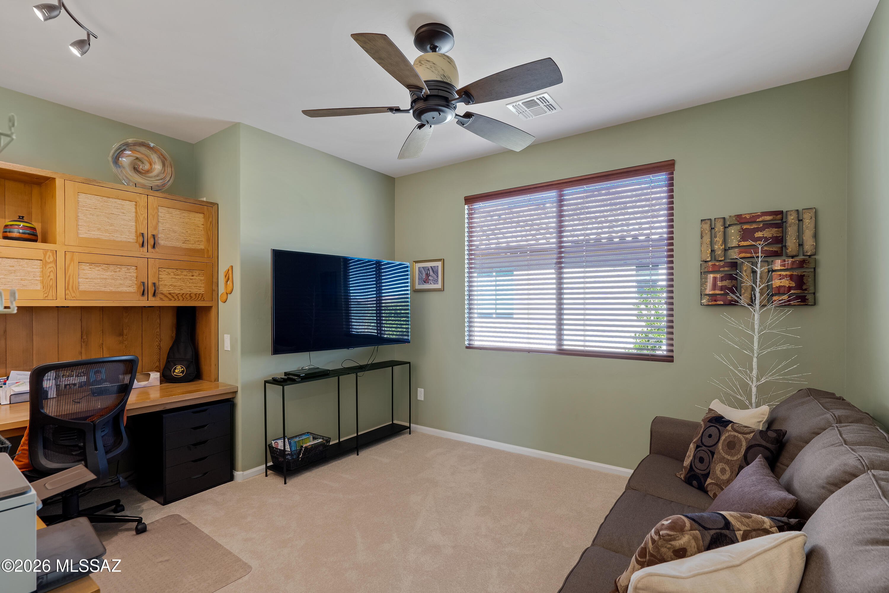 Furnished den with built-in wood desk, television, and gray sofa beside a sunlit window.