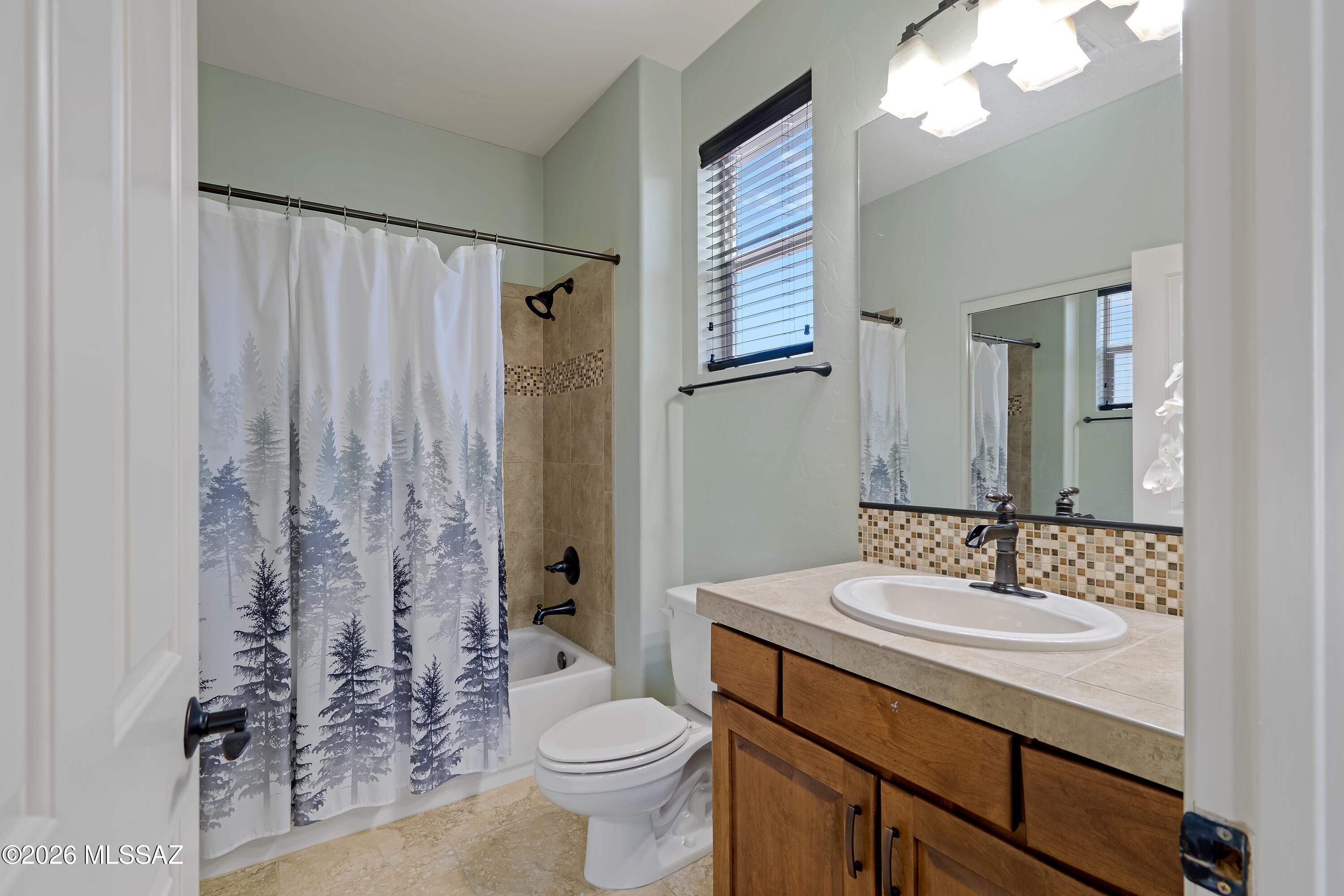 Bathroom with a forest-print shower curtain, tan tiled tub surround, and wood vanity.