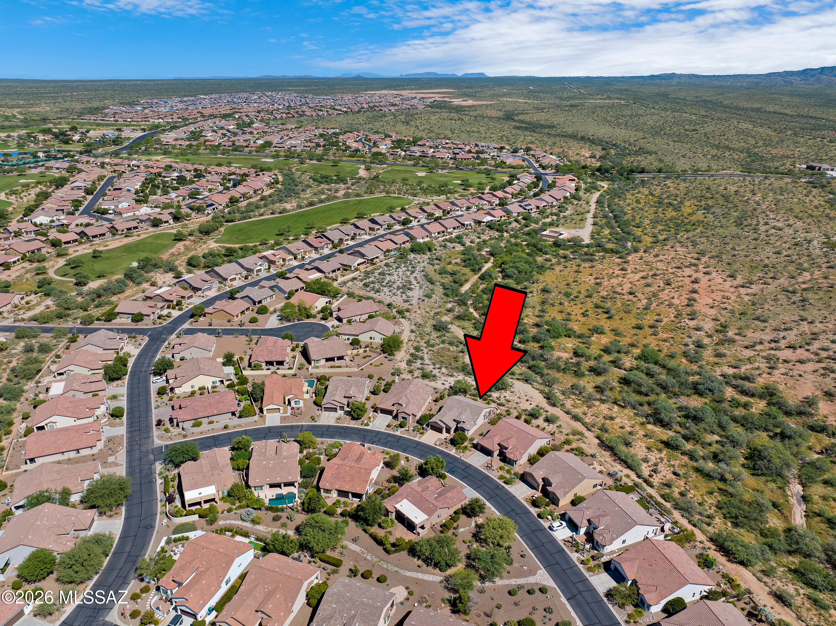Aerial view of a desert subdivision home marked by a red arrow, beside open scrubland.