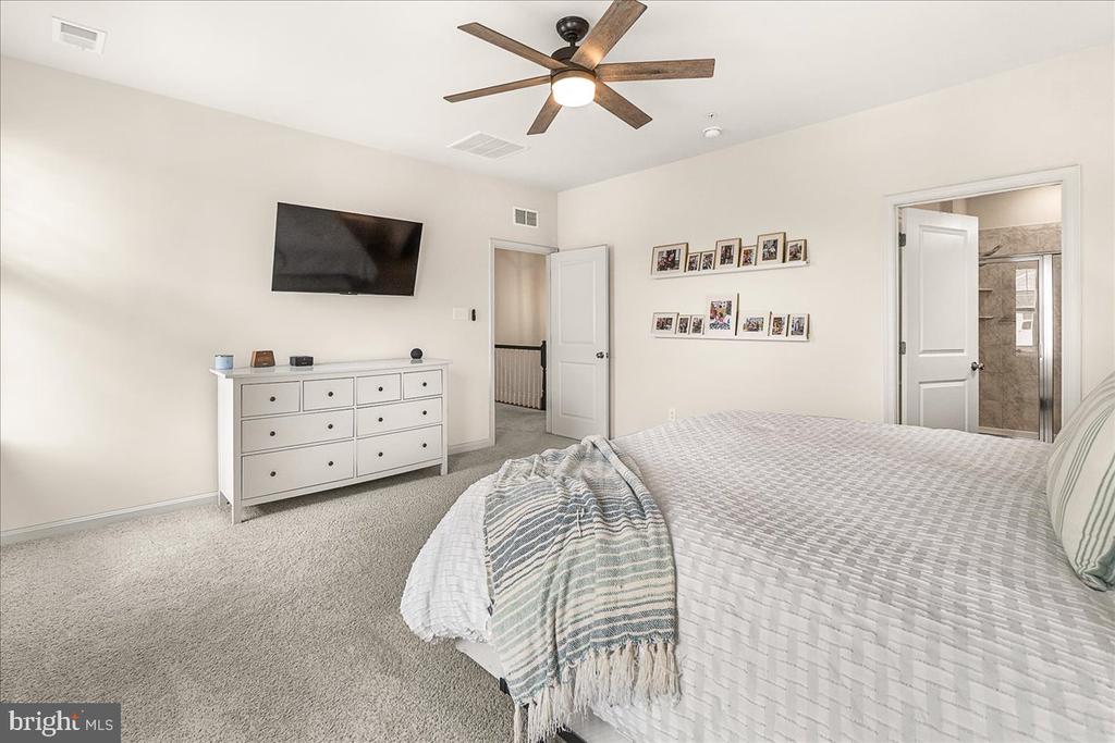Furnished bedroom with a large bed, ceiling fan, wall-mounted television, and doorway to a tiled bathroom.