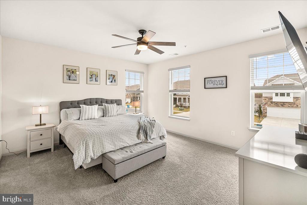 Furnished bedroom with a gray upholstered bed, tufted bench, ceiling fan, and windows overlooking neighboring homes.