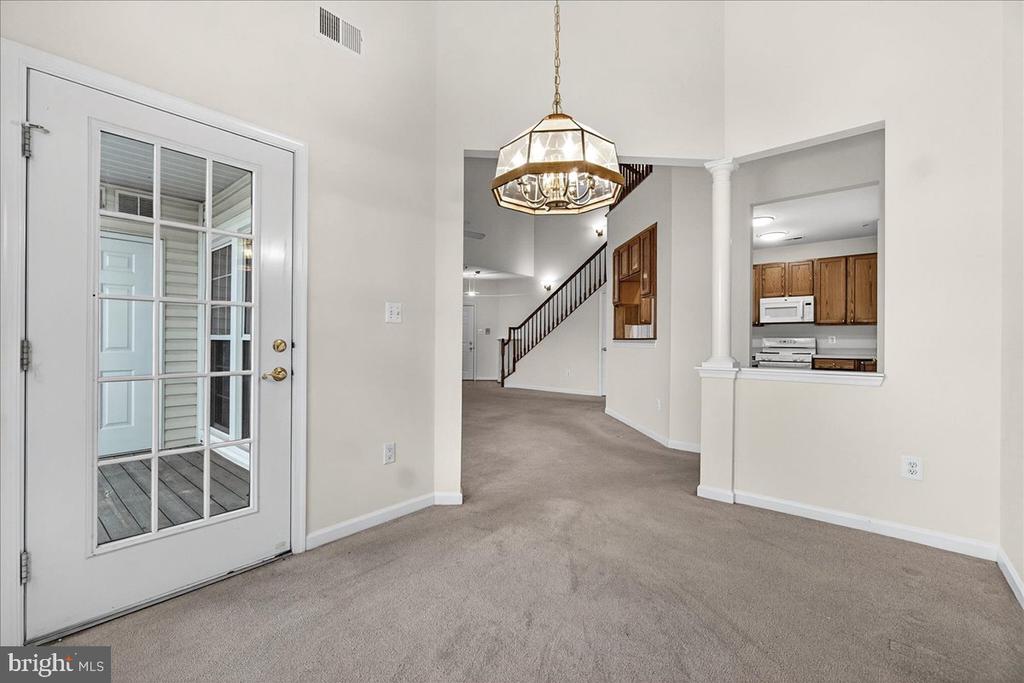 Empty carpeted dining area with a chandelier, glass-paneled door, staircase, and kitchen beyond.