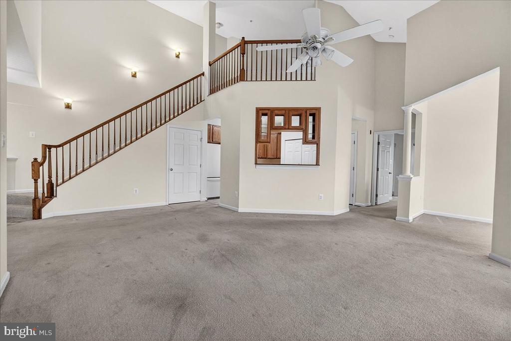 Carpeted living room with a high ceiling, wooden staircase, loft railing, and kitchen pass-through.