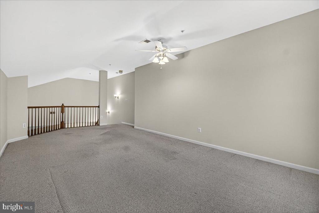Empty carpeted upper-level living area with a ceiling fan and dark wood stair railing.
