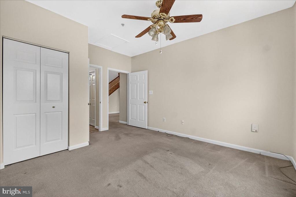 Unfurnished bedroom with beige carpet, white closet doors, and a ceiling fan; staircase visible through the doorway.
