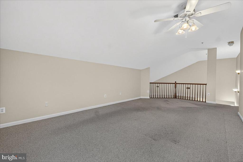 Empty carpeted loft with a ceiling fan and railing overlooking the lower level.