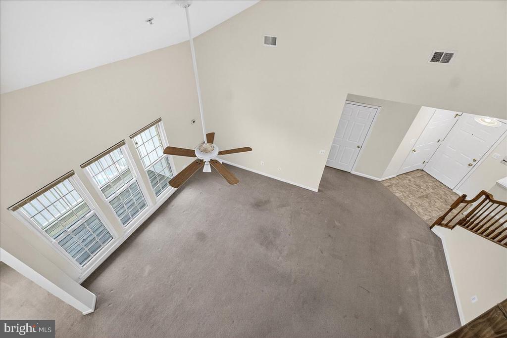 Vacant carpeted living room with a ceiling fan, three tall windows, and a high ceiling.
