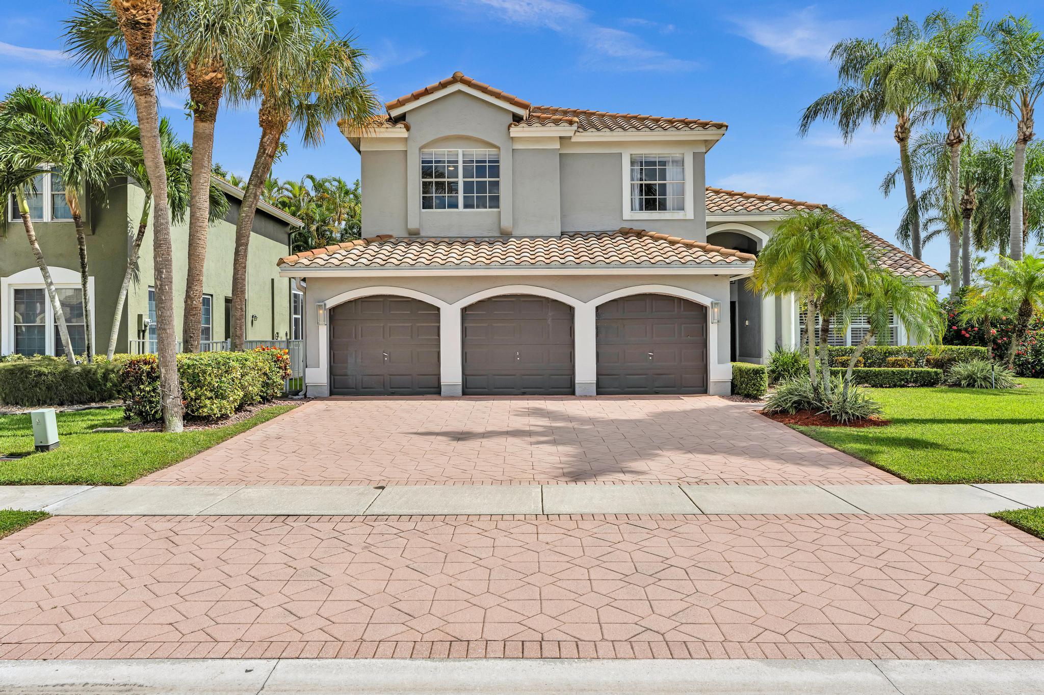Two-story stucco home with a three-car garage, tile roof, palm trees, and a wide paved driveway.