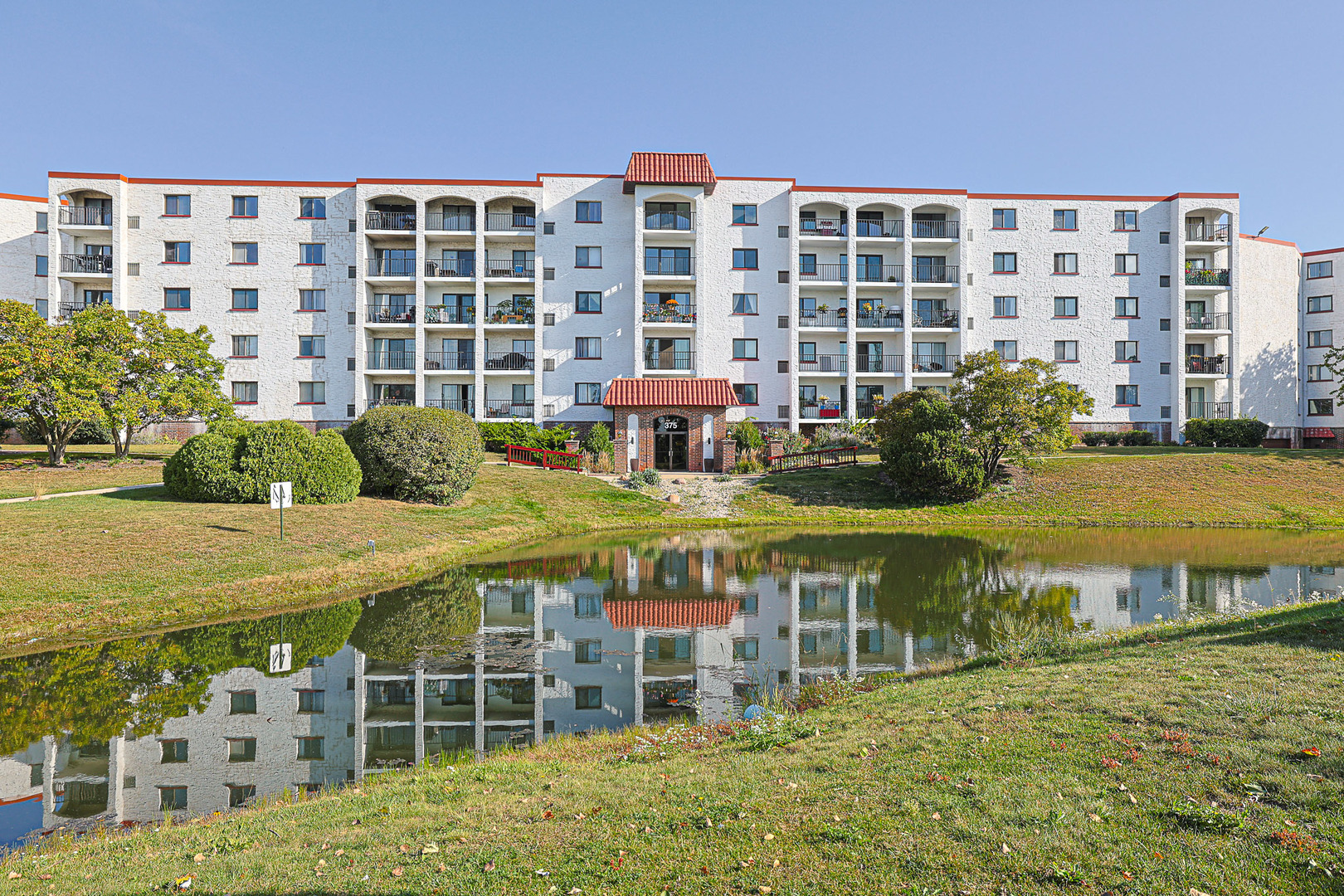 White condominium building with balconies and a red-tile entrance reflected in a pond.