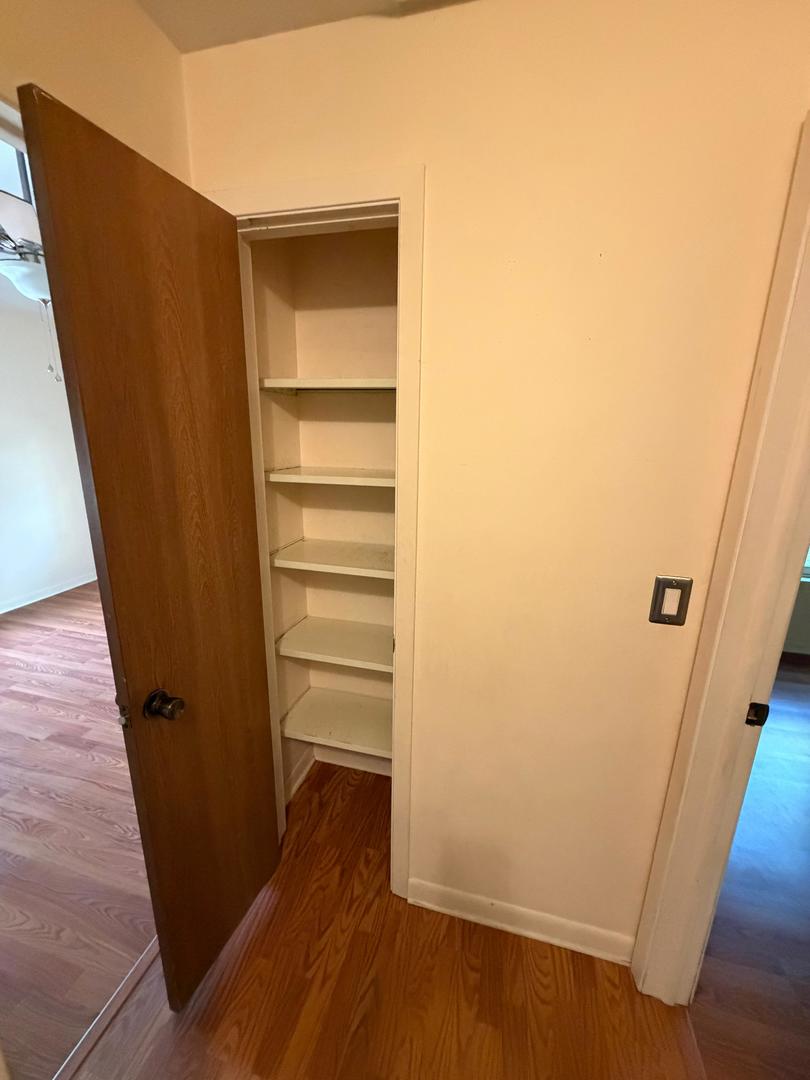 Open narrow closet with built-in shelves beside a wooden door and hardwood flooring.
