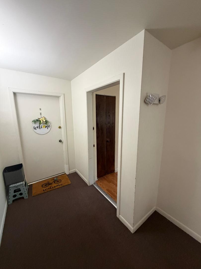 Entry hallway with brown carpet, a welcome-decorated front door, and an open doorway to wood flooring.