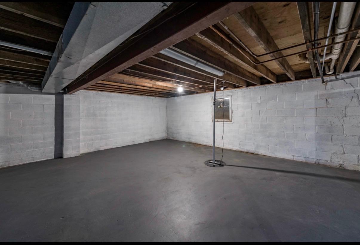 Unfinished basement with an open concrete floor and exposed ceiling beams and pipes.