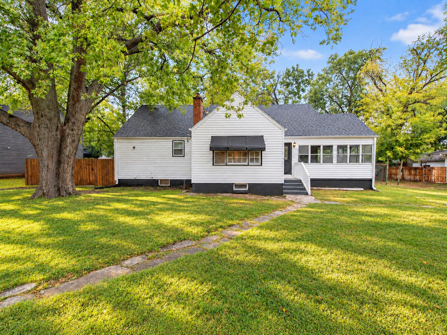 White single-story house with a black window awning and sunroom, set behind a stone path and broad lawn.