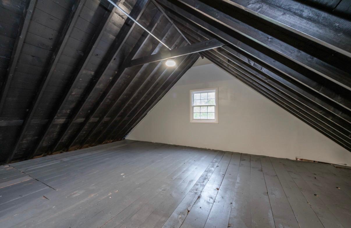 Empty attic with exposed roof rafters, gray plank flooring, and a small window.