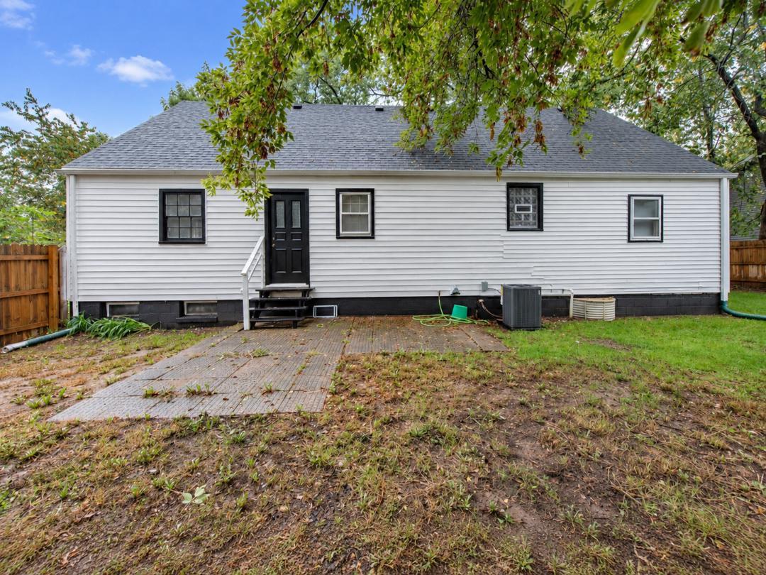 Rear of a white-sided home with a black door and patchy yard beneath leafy trees.
