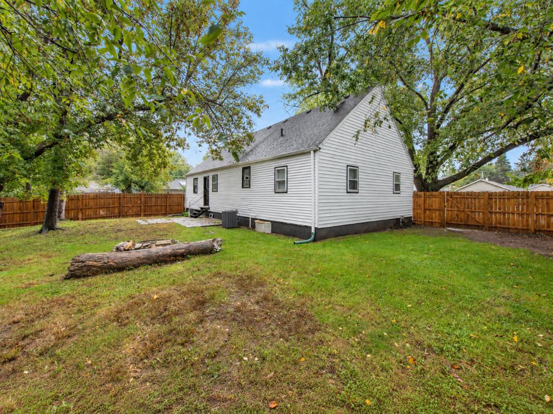 White gable-roofed house beside a fenced backyard with mature trees and a patchy lawn.