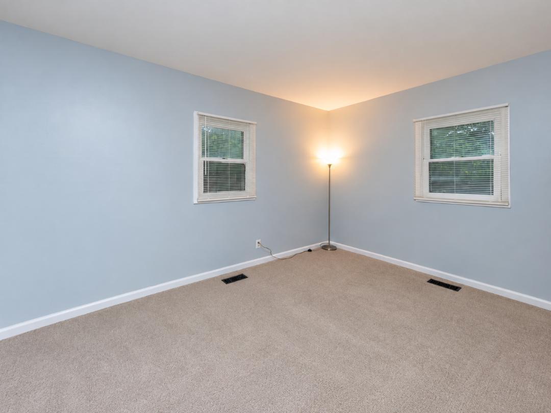 Empty blue-walled room with beige carpet, two windows, and a lit floor lamp.