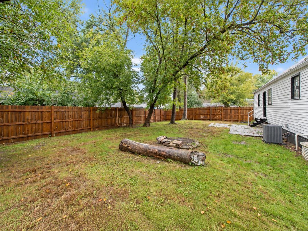 Fenced backyard with mature trees, a patchy lawn, and a fallen log near the house.