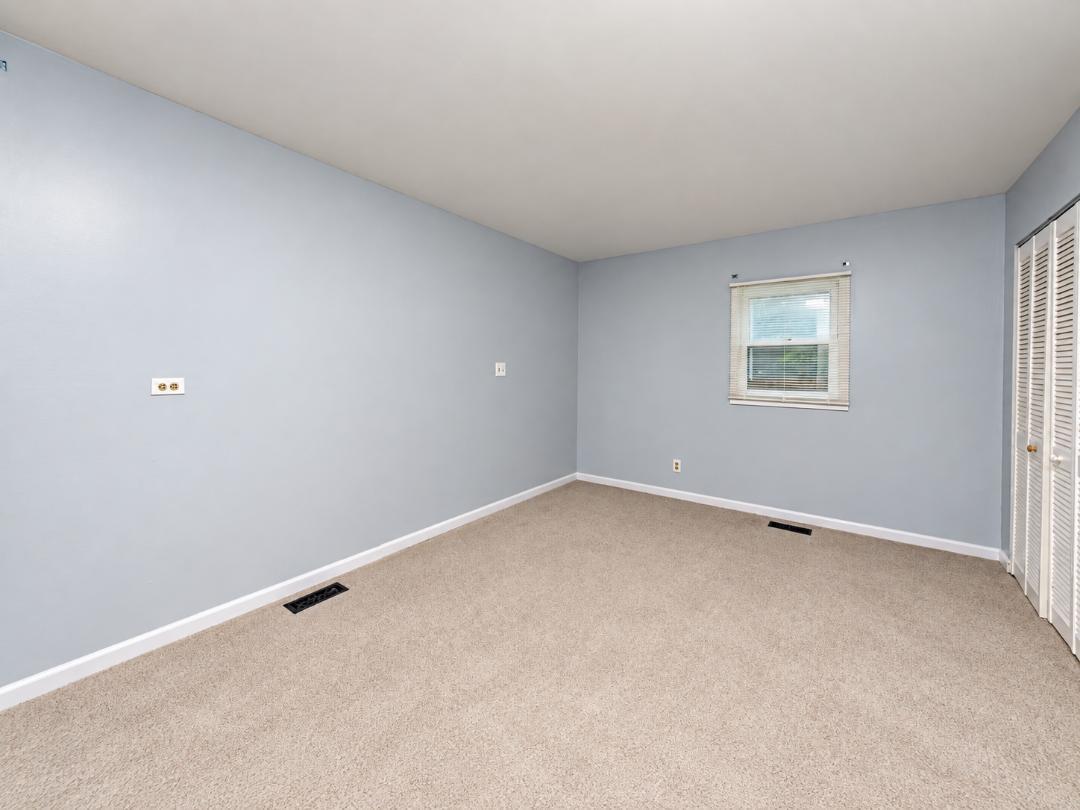Empty bedroom with beige carpet, pale blue walls, a window, and louvered closet doors.