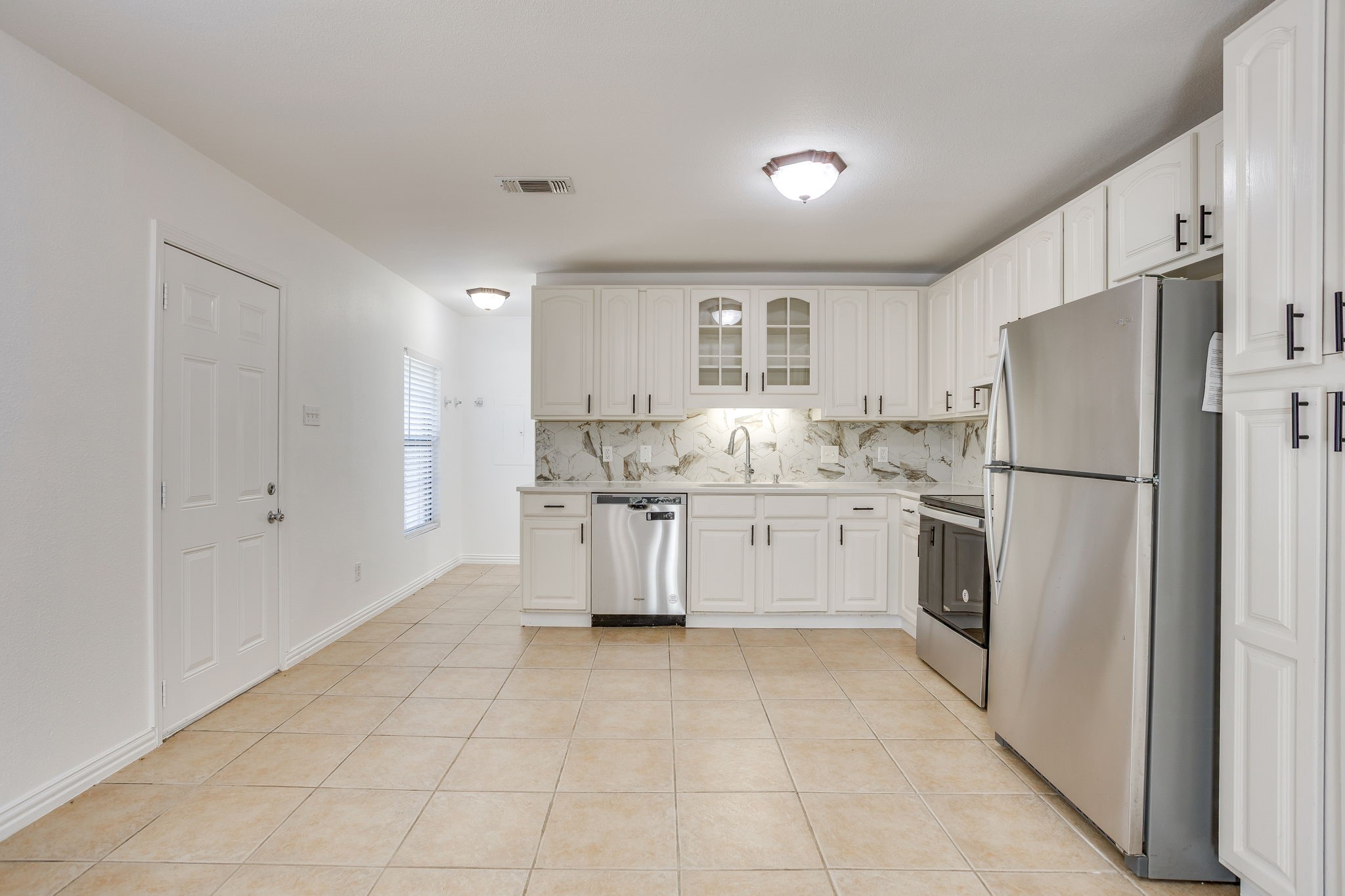 Vacant kitchen with white cabinets, marble-pattern backsplash, tan tile floors, and stainless steel appliances.