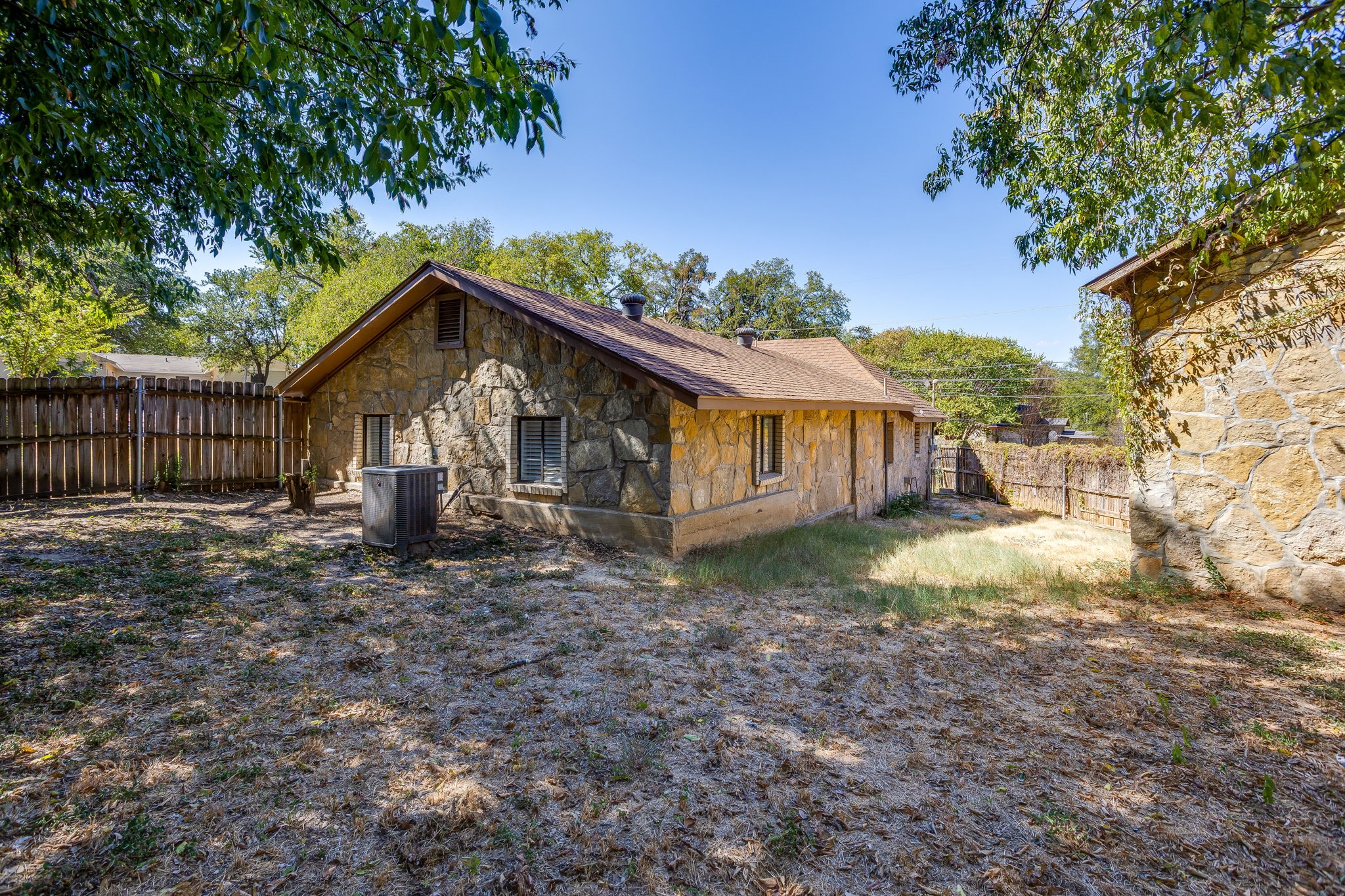 Stone house viewed from the side and rear, with a fenced yard and mature trees.