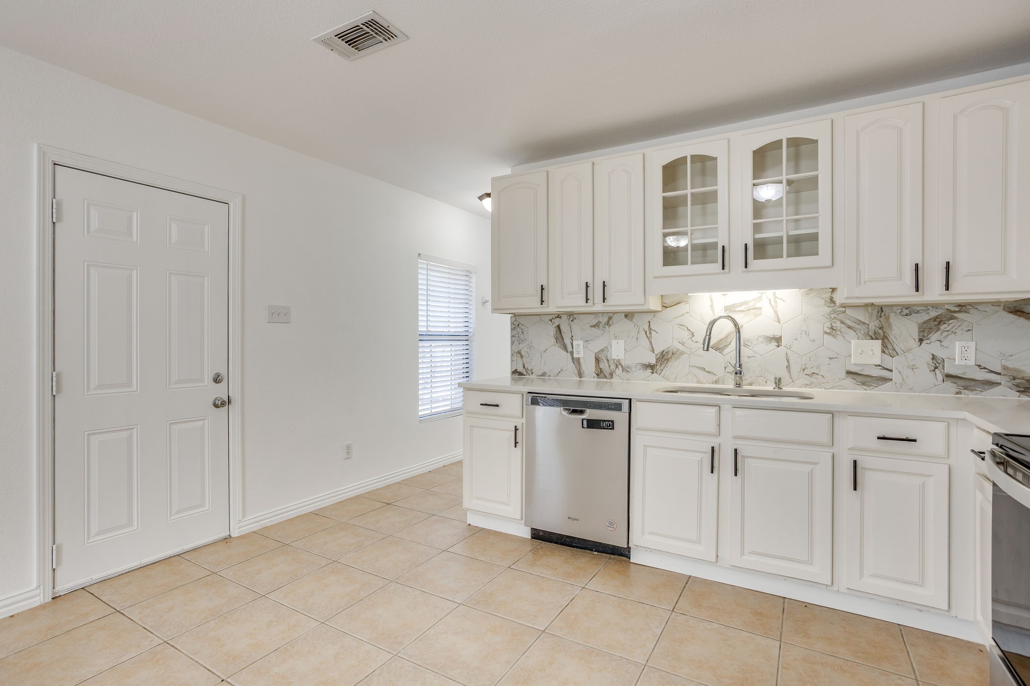 Kitchen with white cabinets, marble-look backsplash, stainless dishwasher, and tan tile floor.