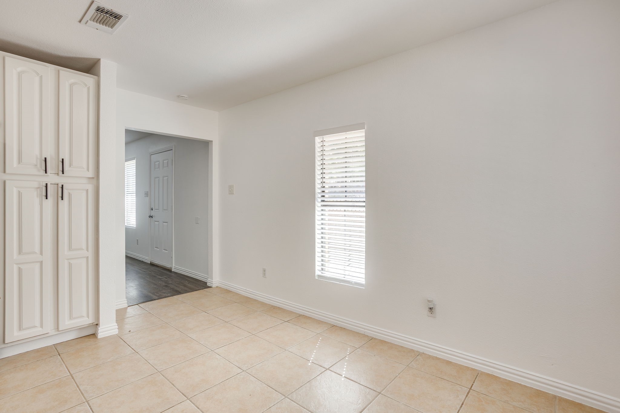 Vacant tiled room with tall white cabinets and a window with blinds beside an open hallway.