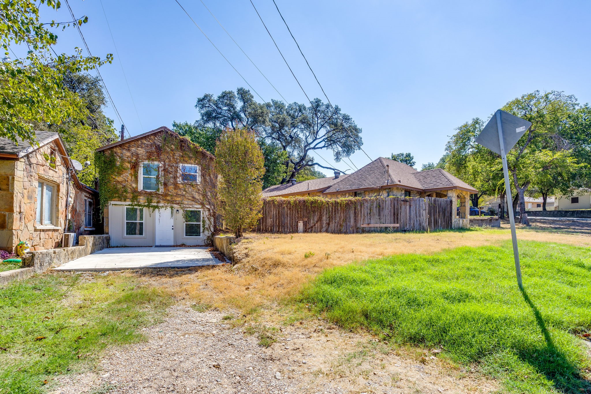 Stone house with a two-story section, gravel driveway, patchy lawn, and wooden fence.