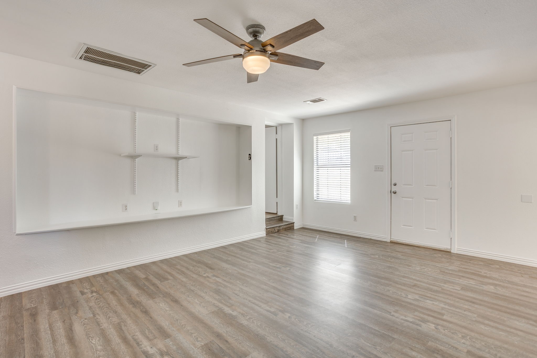 Empty room with plank flooring, built-in shelves, a ceiling fan, and a window beside the door.