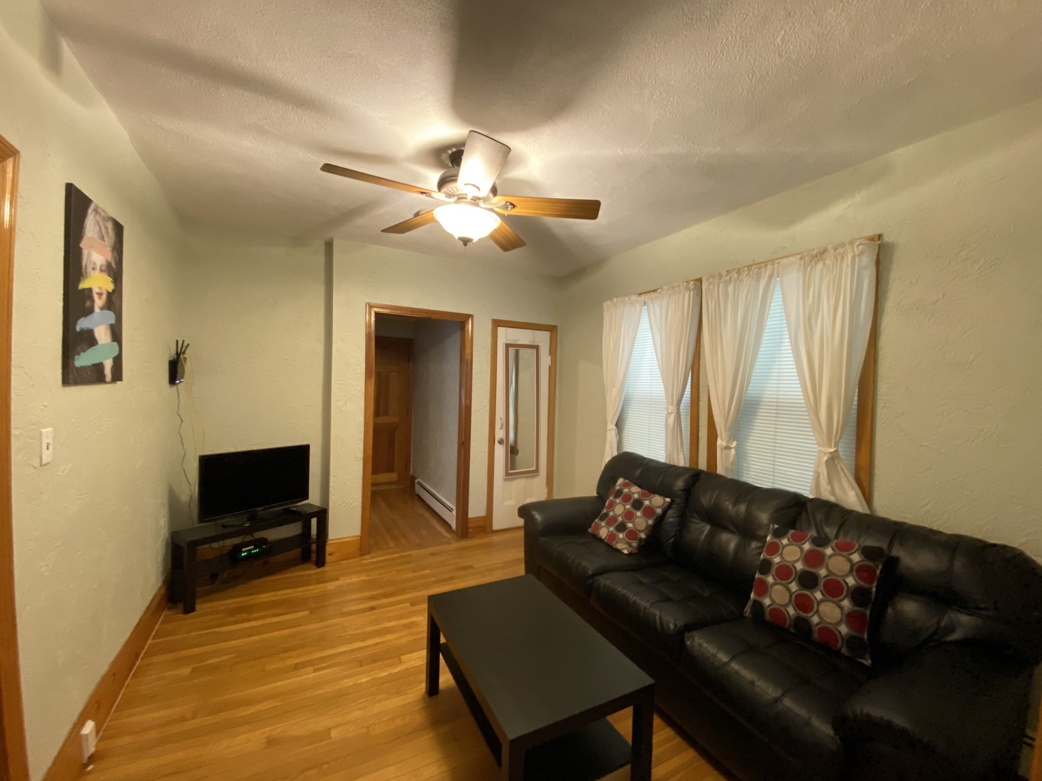 Living room with hardwood floors, a black leather sofa, curtained windows, and a ceiling fan.
