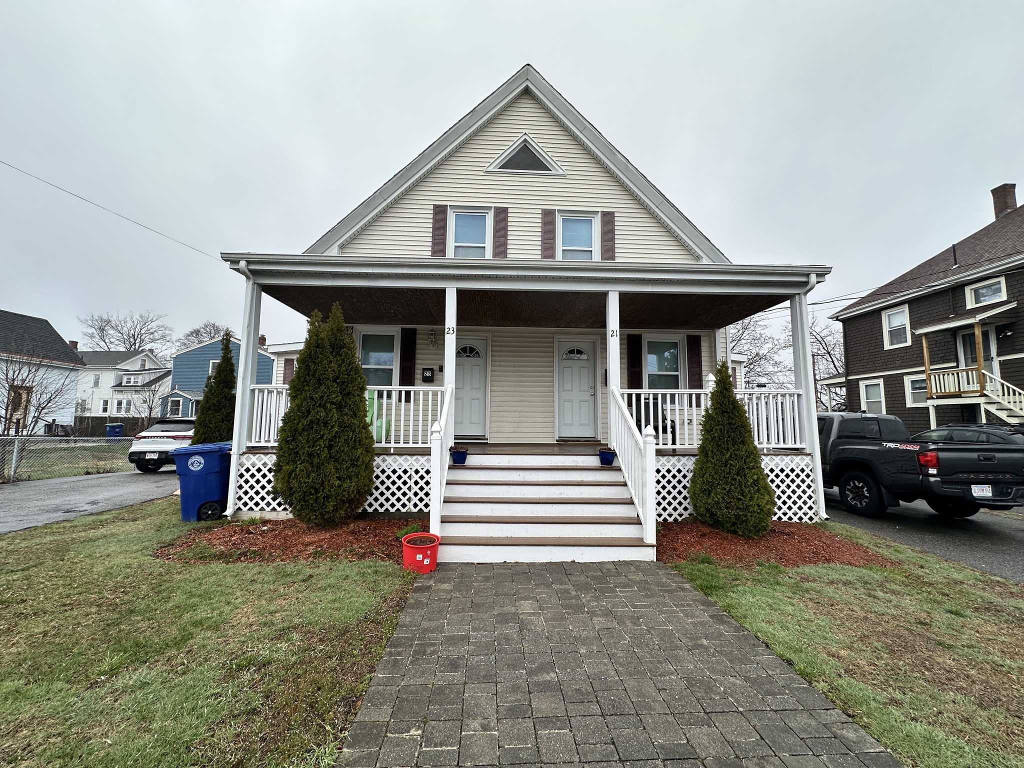 Beige two-unit house with a broad covered porch, white railings, and a paved front walkway.