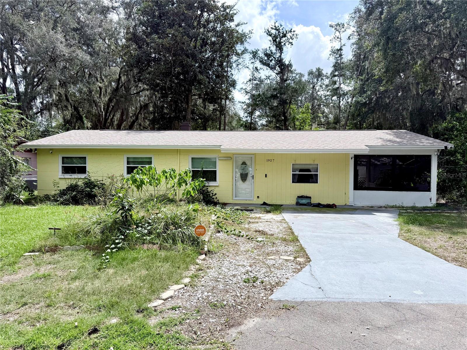 Yellow single-story house with a screened porch, concrete driveway, and moss-draped trees.
