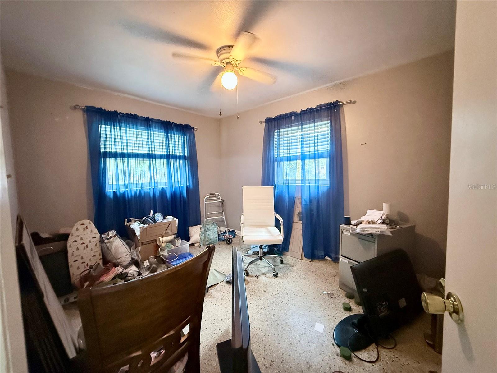 Cluttered room with blue-curtained windows, a ceiling fan, and office furniture.